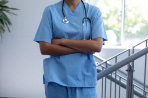 Midsection of female doctor wearing scrubs with stethoscope and arms crossed in hospital corridor
